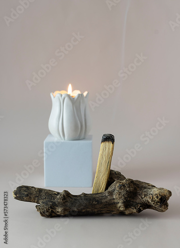 A palo santo wand on a light table. A white candlestick with a tea candle in the background. Light beige background
