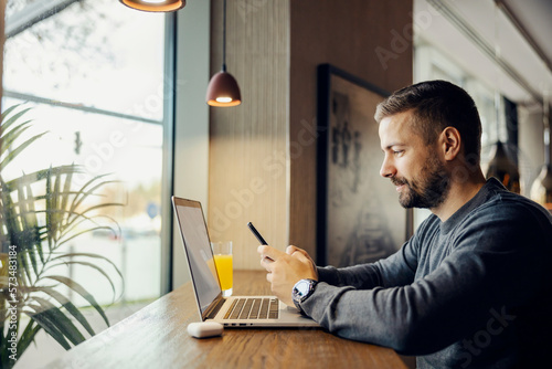 Canvas-taulu A busy digital marketer is sitting in coffee shop and working on social media by using phone and laptop