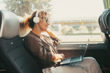 Modern travel lifestyle people with happy smiling woman listening music with headphones and laptop computer sitting and relaxing on a bus seat as a travel passenger. Transportation female people