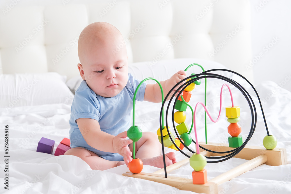 Naklejka premium funny baby boy in blue bodysuit playing with wooden toy on bed