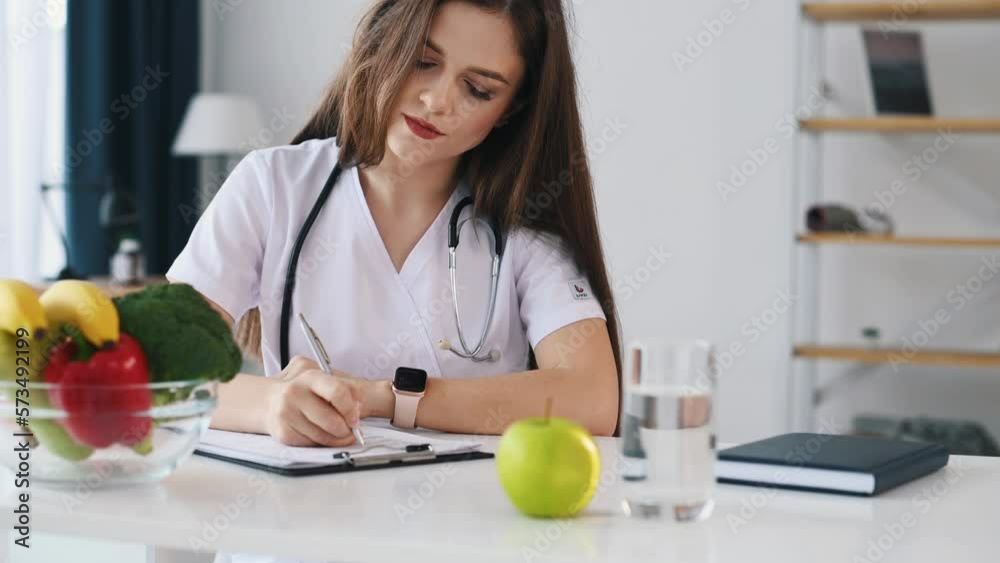 Writing on the documents. Female doctor nutritionist in white uniform is in the clinic. Writing on the documents. Female doctor nutritionist in white uniform is in the clinic.