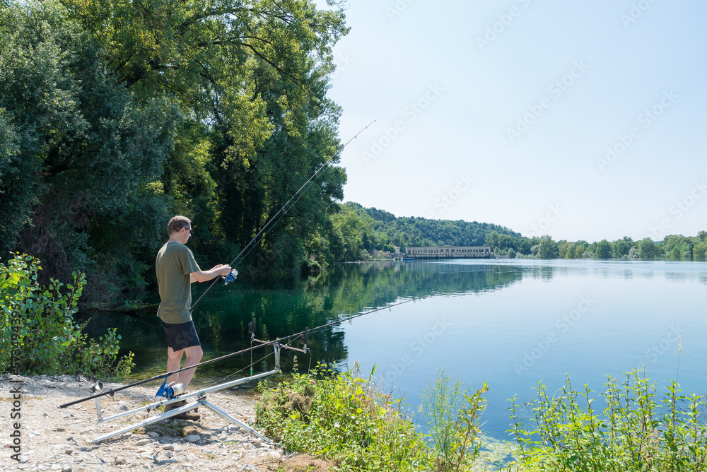 Fishing adventures, carp fishing. Fisherman on the bank of a river with fishing rod in hand and rod pod beside. Outdoor recreation in summer. Ticino river, Italy Fishing adventures, carp fishing. Fisherman on the bank of a river with fishing rod in hand and rod pod beside. Outdoor recreation in summer. Ticino river, Italy