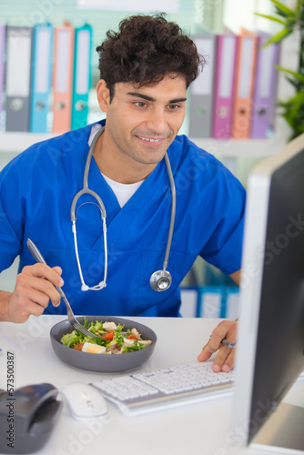 young male doctor having lunch at office using computer photo