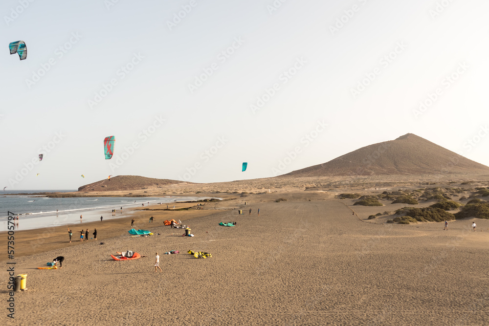 Obraz premium seascape, views of the yellow sand beach, bushes, people practicing kite surfing and Montaña Roja in the background. Paya Grande, El Medano, Tenerife, Canary Islands. Spain