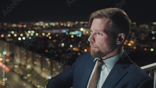 Close-up tilting shot of hands and face of young businessman in suit and tie standing on top floor of office building late in evening, typing message on smartphone, looking away and sighing ruefully photo
