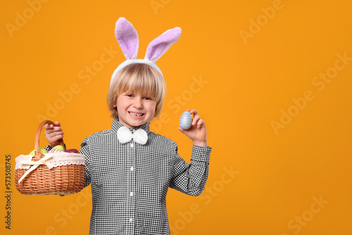 Happy boy in bunny ears headband holding wicker basket with painted Easter eggs on orange background. Space for text