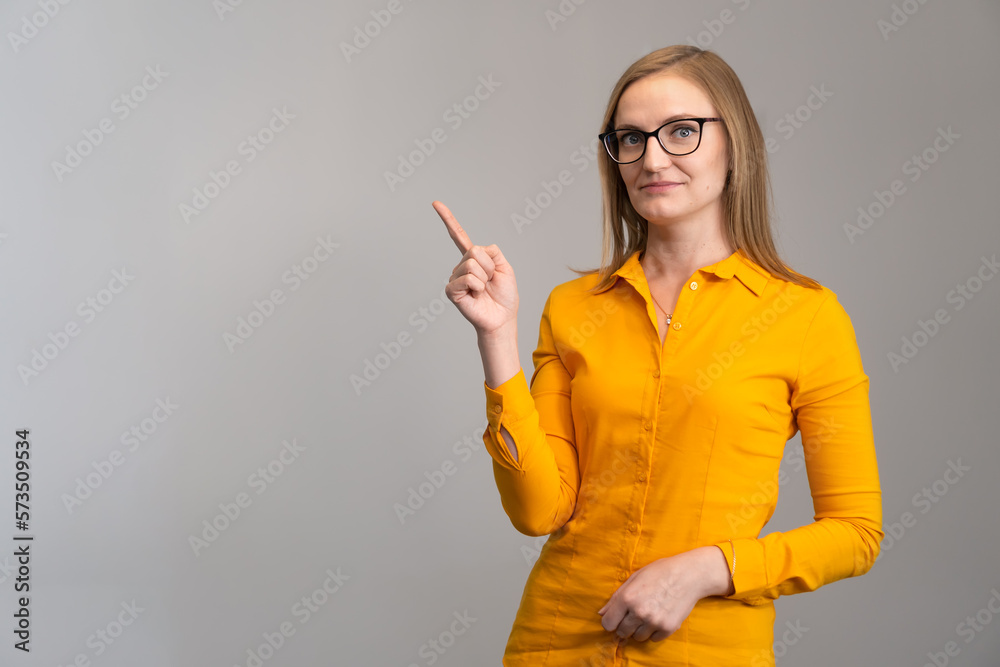 Woman in glasses and an orange shirt points her finger at an empty seat on a light background. Concept of advertising, business, training Woman in glasses and an orange shirt points her finger at an empty seat on a light background. Concept of advertising, business, training