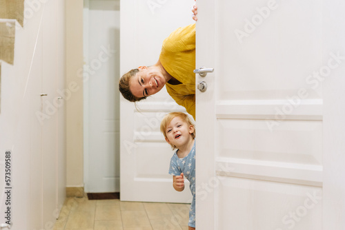 A young mother and little daughter play hide-and-seek behind the door of the house.