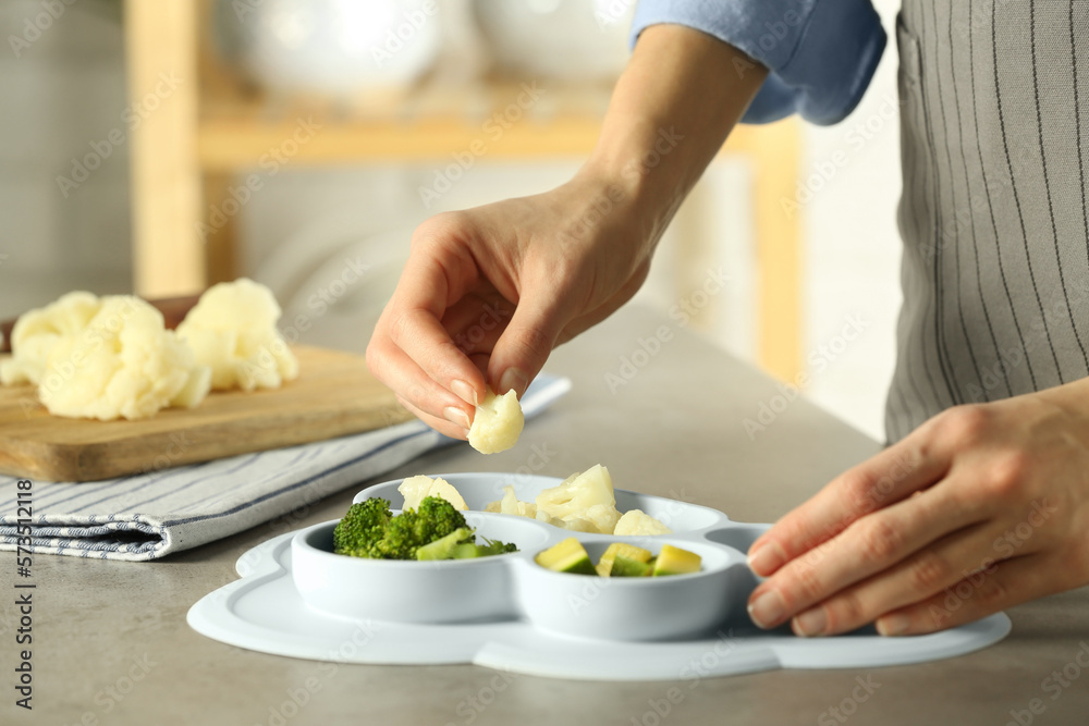 Baby food. Woman putting piece of cauliflower into section plate with different vegetables at grey textured table in kitchen, closeup