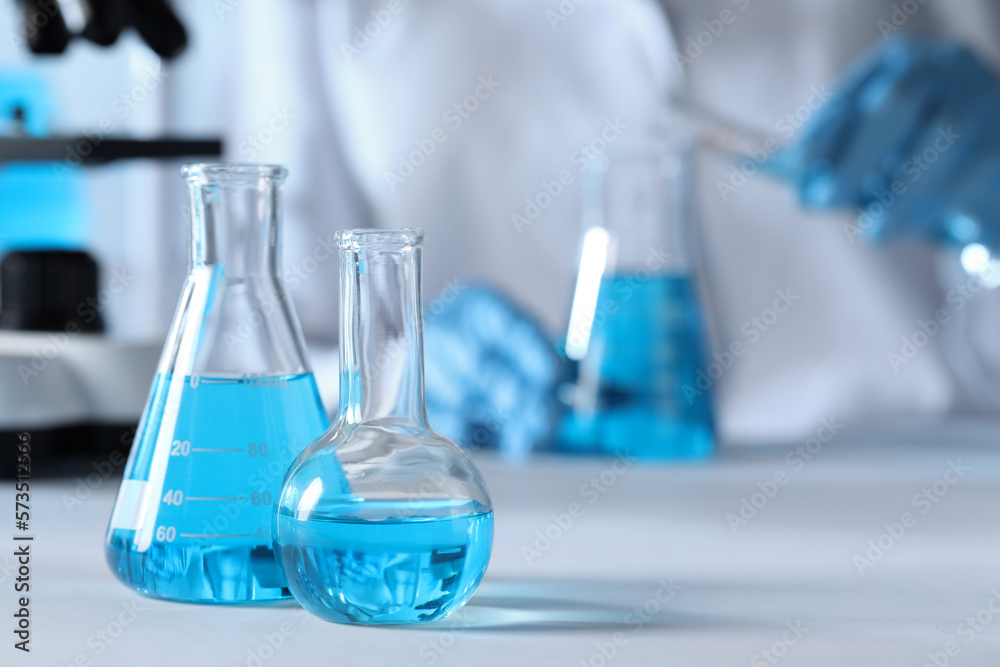 Closeup view of scientist pouring light blue liquid from test tube into flask and laboratory glassware on table, selective focus Closeup view of scientist pouring light blue liquid from test tube into flask and laboratory glassware on table, selective focus