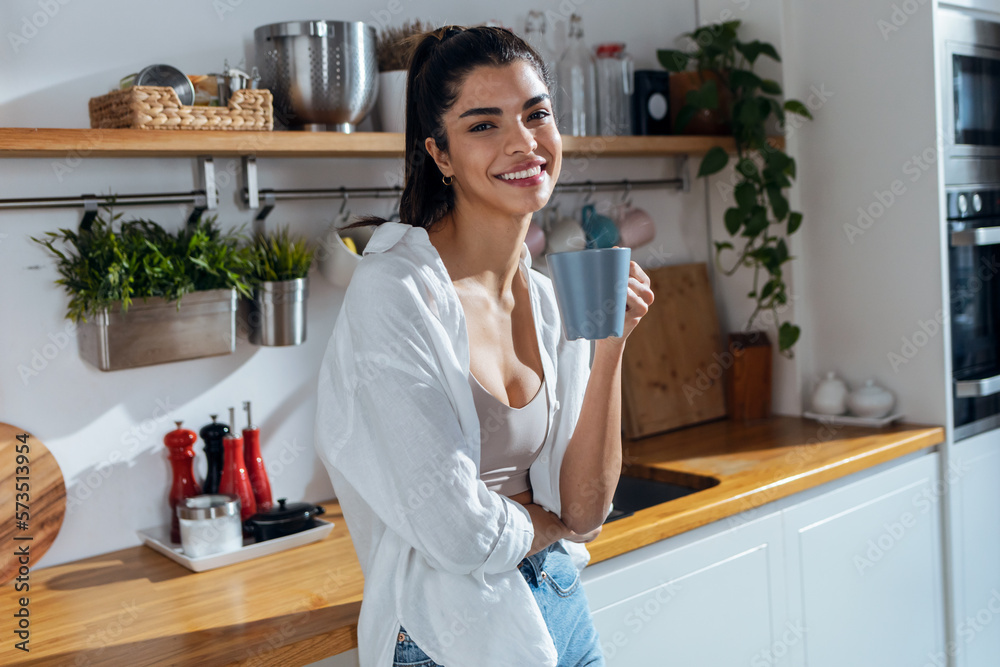 Beautiful young woman drinking coffee while standing in the kitchen at morning