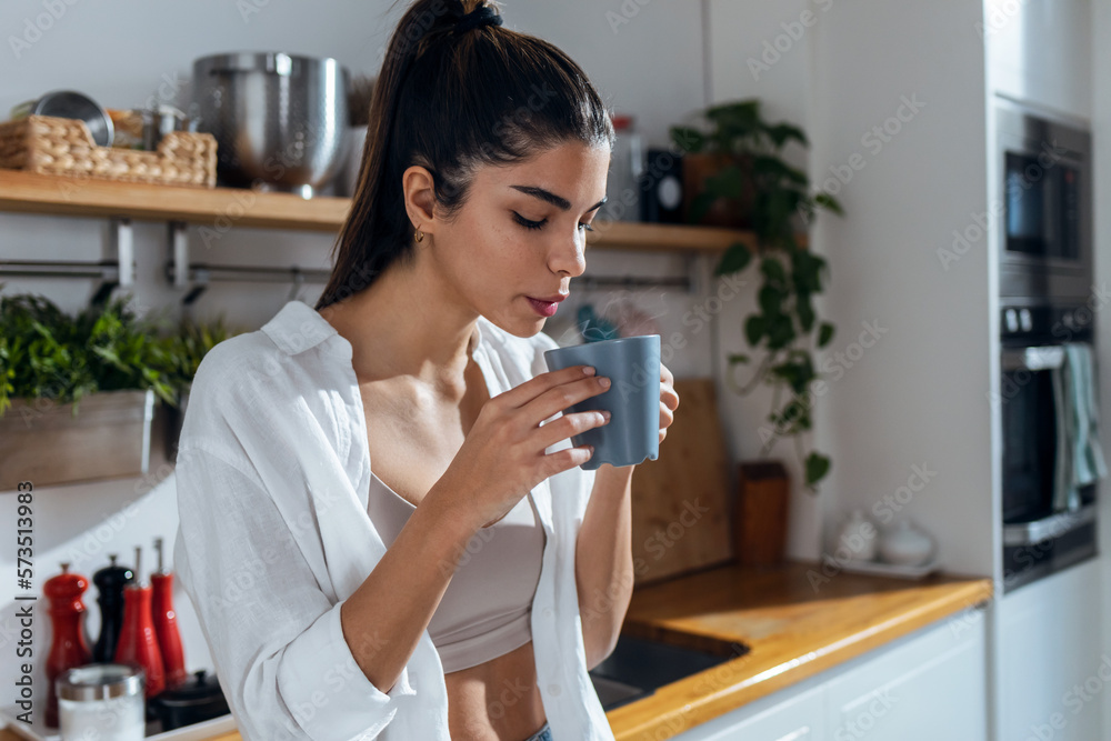 Beautiful young woman drinking coffee while standing in the kitchen at morning Beautiful young woman drinking coffee while standing in the kitchen at morning