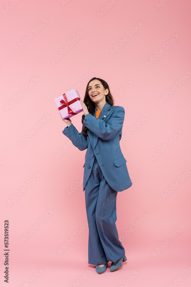 full length of happy woman in blue pantsuit holding wrapped present with ribbon on pink. full length of happy woman in blue pantsuit holding wrapped present with ribbon on pink.