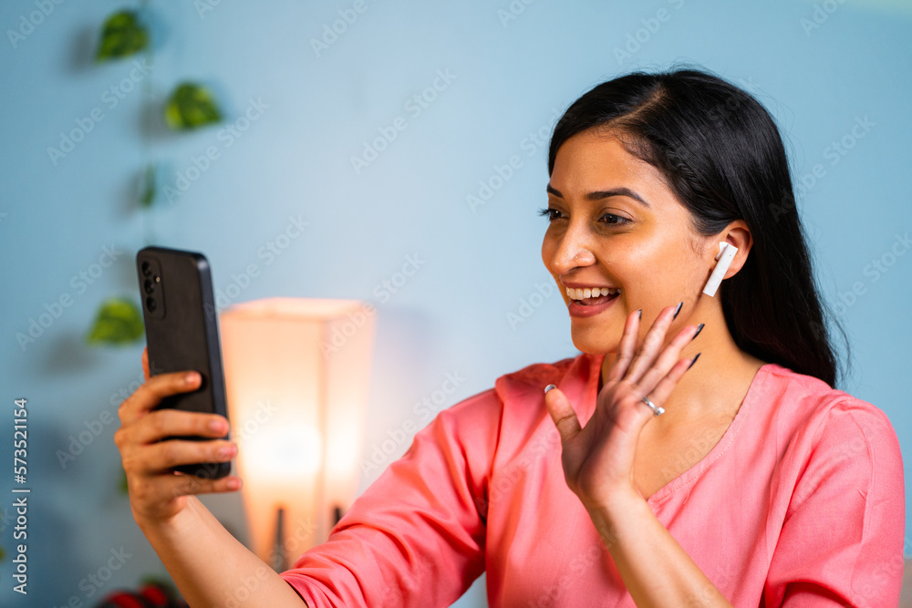 Happy young girl with earbuds talking in video call on mobile phone at home - concept of technology, connection and communication. Happy young girl with earbuds talking in video call on mobile phone at home - concept of technology, connection and communication.