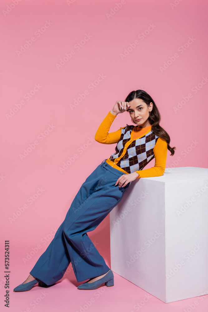 Fototapeta premium full length of pretty woman in trendy cardigan and blue pants leaning on white cube on pink background.