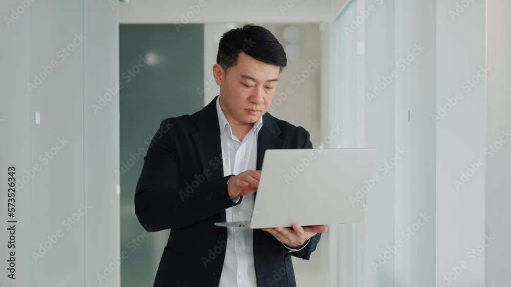 Handsome korean man working on laptop project in office hallway. Online business negotiations. Serious businessman chatting with foreign partners, writing message, look to window, thinking new idea. Handsome korean man working on laptop project in office hallway. Online business negotiations. Serious businessman chatting with foreign partners, writing message, look to window, thinking new idea.