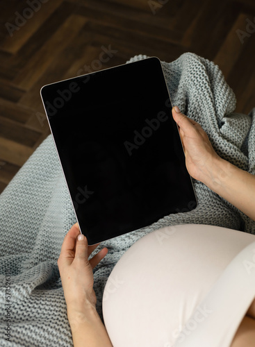 A pregnant girl with a large belly holds a touchscreen tablet in her hands. A pregnant woman makes purchases in an online store, talks via video link with relatives or a doctor. Photo of a pregnant