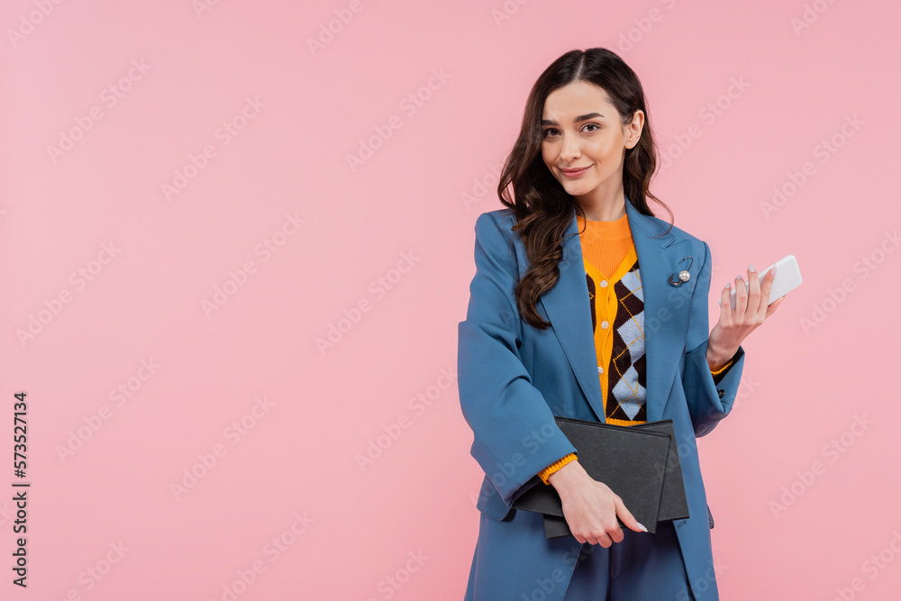 cheerful young woman in blue blazer holding smartphone and notebooks isolated on pink. cheerful young woman in blue blazer holding smartphone and notebooks isolated on pink.