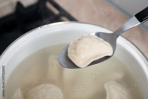 Spoon with tasty dumpling (varenyk) over pot indoors, closeup