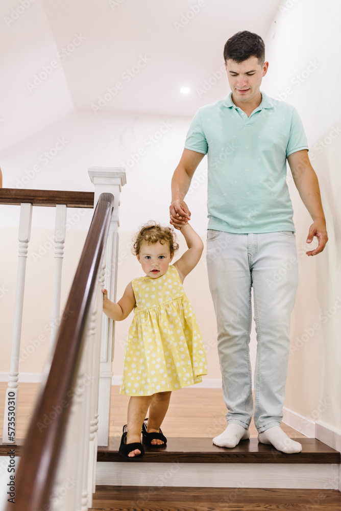 Fototapeta premium Dad holds his daughter's hand and leads her up the stairs. A girl put on her mother's shoes with father on stairs at home. Dangerous situation at home. Kid walking stairs. Baby playing in staircase.