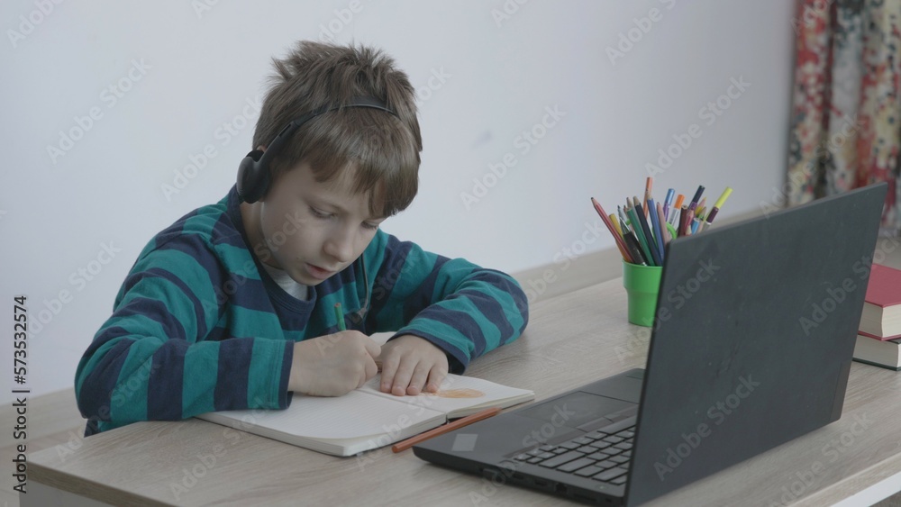 Portrait of boy with headphone sit at desk and write, boy look at open laptop