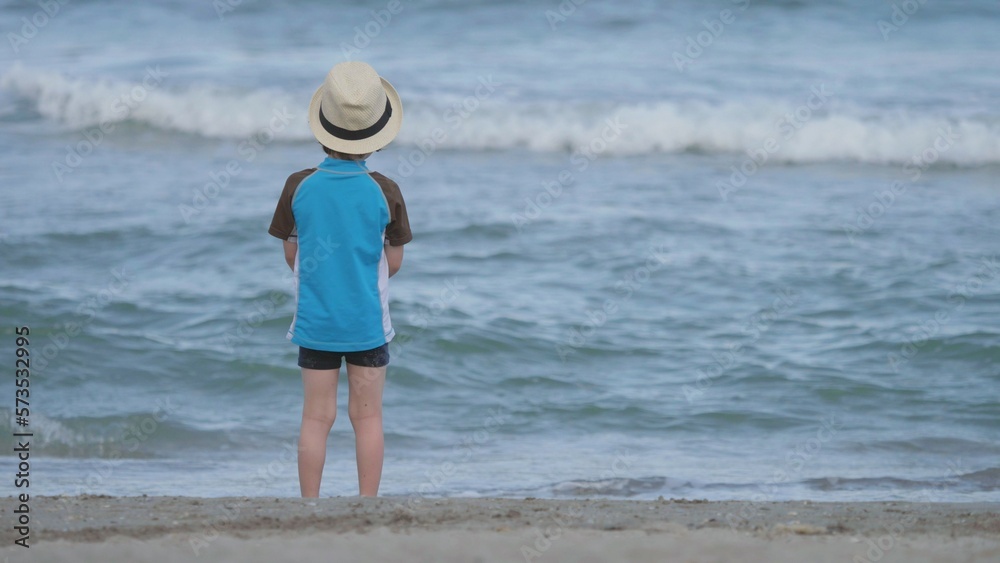 Isolated little child with had on beach throw of sand in blue sea water
