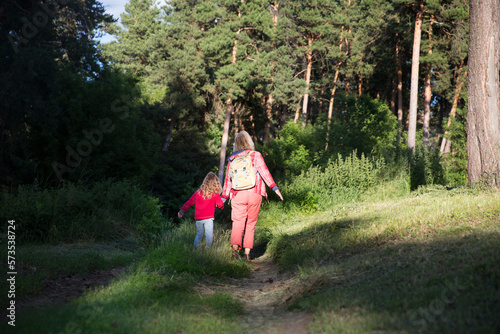 mom and and child daughter with backpack back to the viewer walking in the forest. Staycations, hyper-local travel, family outing, getaway, natural environ. Concept of friendly family. 