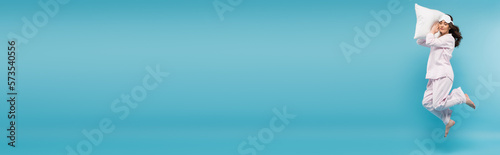 full length of barefoot young woman in night mask and sleepwear holding pillow and levitating on blue background, banner.