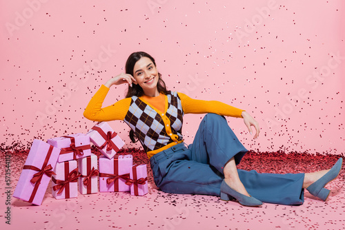 smiling woman in stylish attire sitting near pile of gift boxes and festive confetti on pink background.