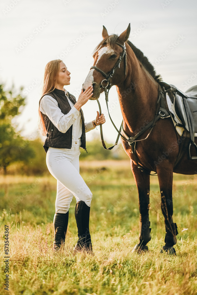 Obraz premium Young beautiful female jockey strokes and hugs the horse's head and prepares for the competition. Jumping training in the meadow in summer evening.