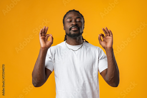 Concentrated relaxed african man standing with closed eyes against yellow background