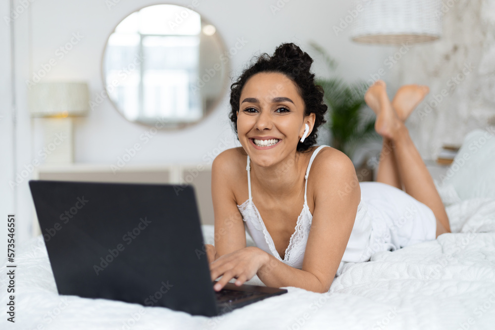 Happy millennial caucasian brunette lady in wireless headphones lies on soft bed, typing on computer Happy millennial caucasian brunette lady in wireless headphones lies on soft bed, typing on computer