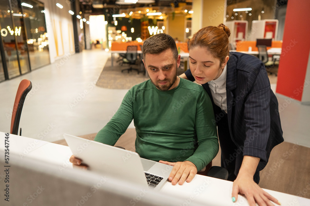 A red-haired Caucasian woman stands at the desktop of a bearded man. The boss approves the work of the subordinate. A red-haired Caucasian woman stands at the desktop of a bearded man. The boss approves the work of the subordinate.