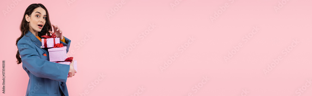 astonished woman in blue blazer holding gift boxes and looking at camera isolated on pink, banner.