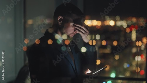 Medium tilting down shot of businessman in suit standing in corporate office late at night, typing on smartphone, rubbing forehead, shaking head, with bokeh lights and female colleague in background photo