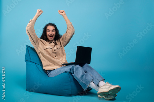 Laughing shocked excited millennial european female student sitting on bag chair with laptop, raising hands up © Prostock-studio