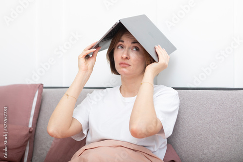 Upset and tired woman lying in the bed and holding laptop over her head