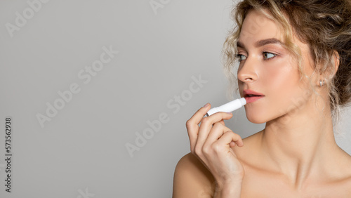 Young beautiful woman with perfect skin applying hygenic lipbalm on lips, looking aside at free space, grey background photo