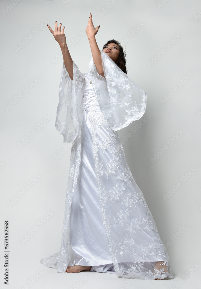 Naklejka premium Full length portrait of beautiful woman wearing fantasy costume, white bridal gown. Standing pose with low angle perspective, gestural arm movements casting a spell. Isolated white studio backgroun