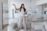 Cheerful brunette young adult woman in white t-shirt and beige pants standing on cozy sofa holds tablet looks at camera. Purposeful  caucasian woman relaxing at hotel. Real estate, mortgage.