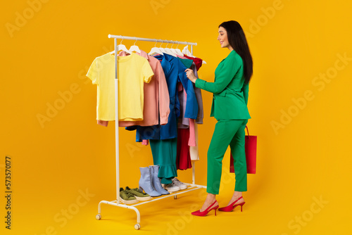 Young female shopaholic shopping and picking outfit, choosing colorful clothes on rail, standing over yellow background