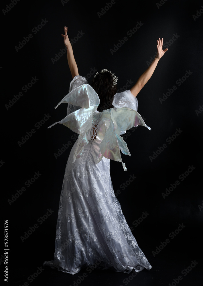 portrait of beautiful woman wearing fantasy costume with white bridal gown and translucent fairy wings. Posing with gestural arms reaching out, isolated on black studio background. portrait of beautiful woman wearing fantasy costume with white bridal gown and translucent fairy wings. Posing with gestural arms reaching out, isolated on black studio background.