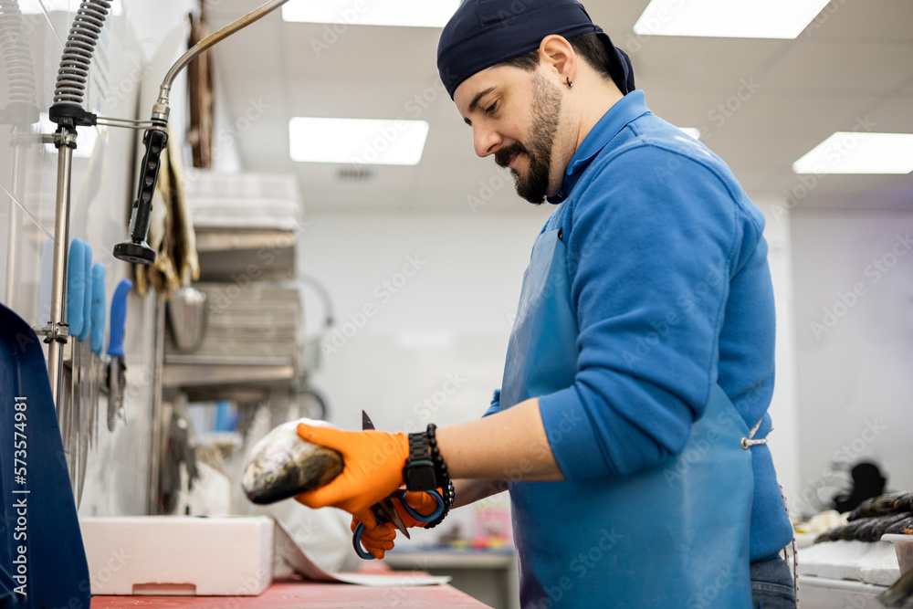 A middle-aged fishmonger in apron and hat is cutting the parts of a fish to clean it with scissors, food sales, small and medium business concept. A middle-aged fishmonger in apron and hat is cutting the parts of a fish to clean it with scissors, food sales, small and medium business concept.