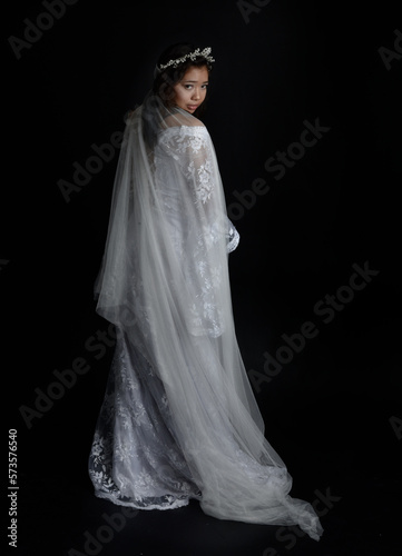 Full length portrait of beautiful woman wearing white bridal fantasy gown with butterfly mask and ethereal veil. Dancing with gestural arm poses, isolated on black studio background.