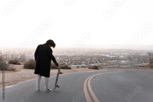 A young man wearing a gabardine is posing with his skate during the sunset in Baldwin Hills, Los Angeles photo