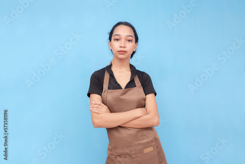 A young asian female barista looking serious with her arms crossed. Wearing a brown bib apron and black shirt and hair let down. Against a light blue background.