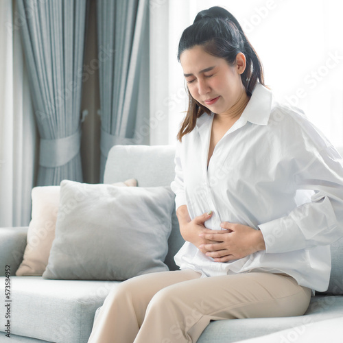 Woman suffering with severe stomach pain sitting at home. Woman's hand holding stomach suffering from stomachache, diarrhea or indigestion problem. Young woman pressing on belly on painful sensation.