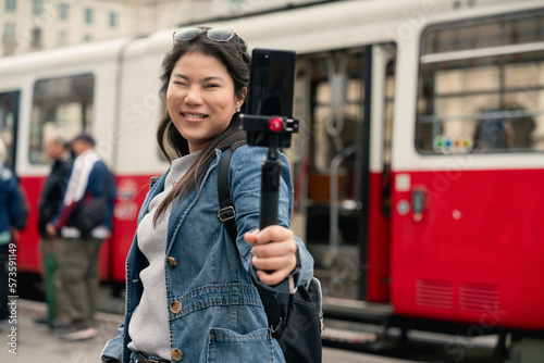 Young casual woman using smartphone to record journey with smile. Happy and excited asian girl traveler wear stylish cloth with hand gesture waving to internet audience. Influencer vlogging concept