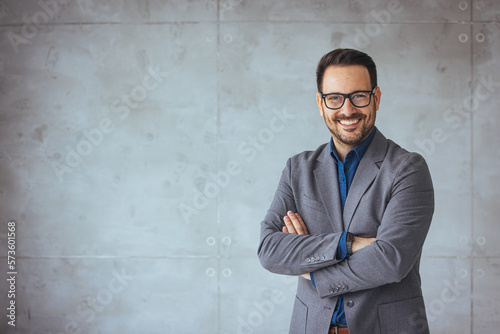 Portrait of happy businessman with arms crossed standing in office. Portrait of young happy businessman wearing grey suit and blue shirt standing in his office