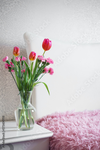 a bouquet of spring pink flowers in a glass vase on a nightstand near a white bed with a pink bedspread. freshness and lightness in the interior